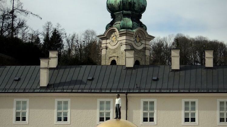 Photo by Heinz Klier A man standing on a golden sphere sculpture in a city square with a church tower in Salzburg, Austria.