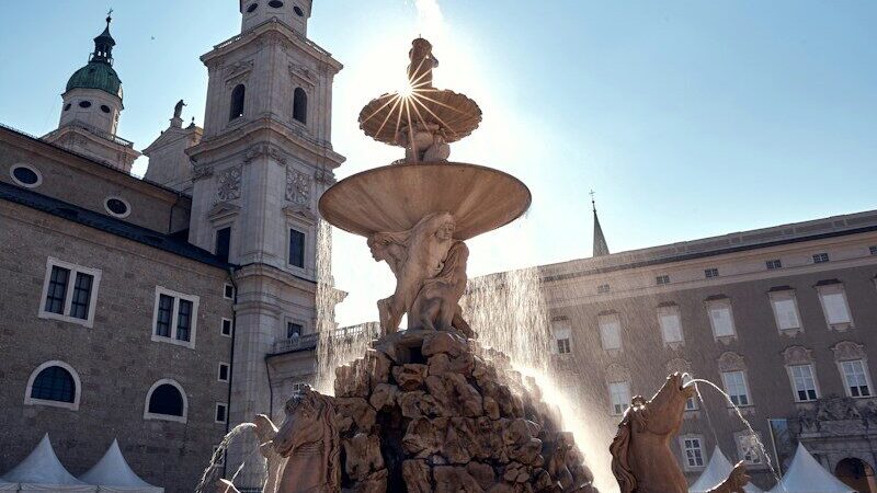Photo by Timo Trilk a fountain in front of a building with a clock tower in the background